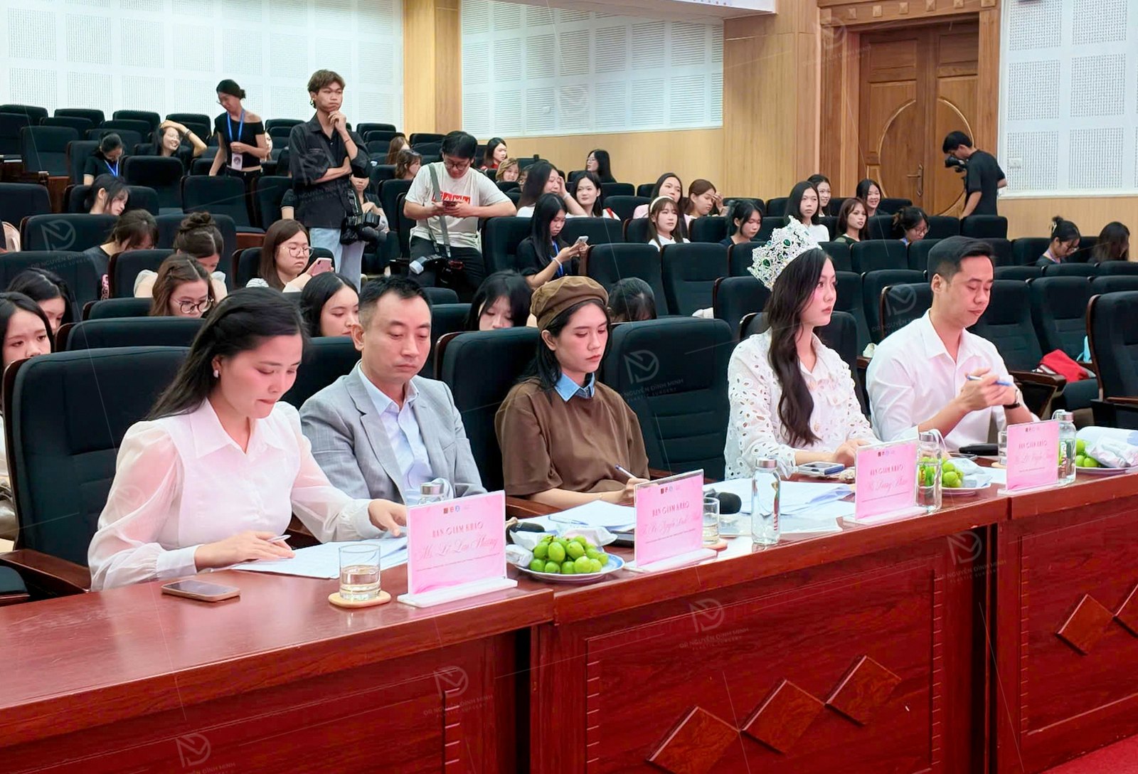 Bốn thành viên ban giám khảo ngồi tại bàn chấm trong hội trường, mỗi người có một biển tên, phía sau là sinh viên dự thi
