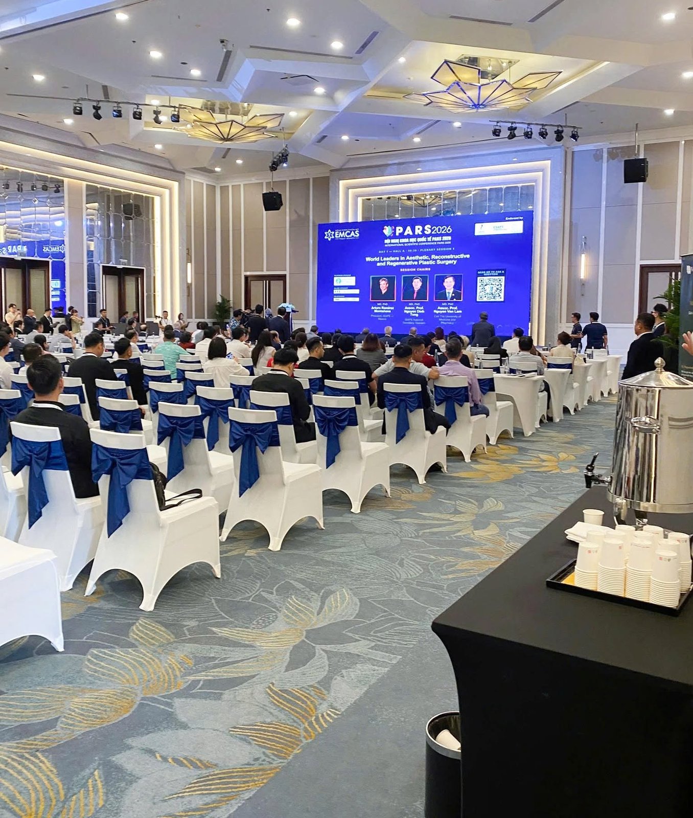 Hội trường phiên toàn thể của hội nghị PARS 2026 tại Hà Nội nhìn từ phía sau: đại biểu ngồi theo các dãy ghế bọc trắng thắt nơ xanh, màn hình lớn ghi PARS 2026 và tên ba chủ tọa phiên