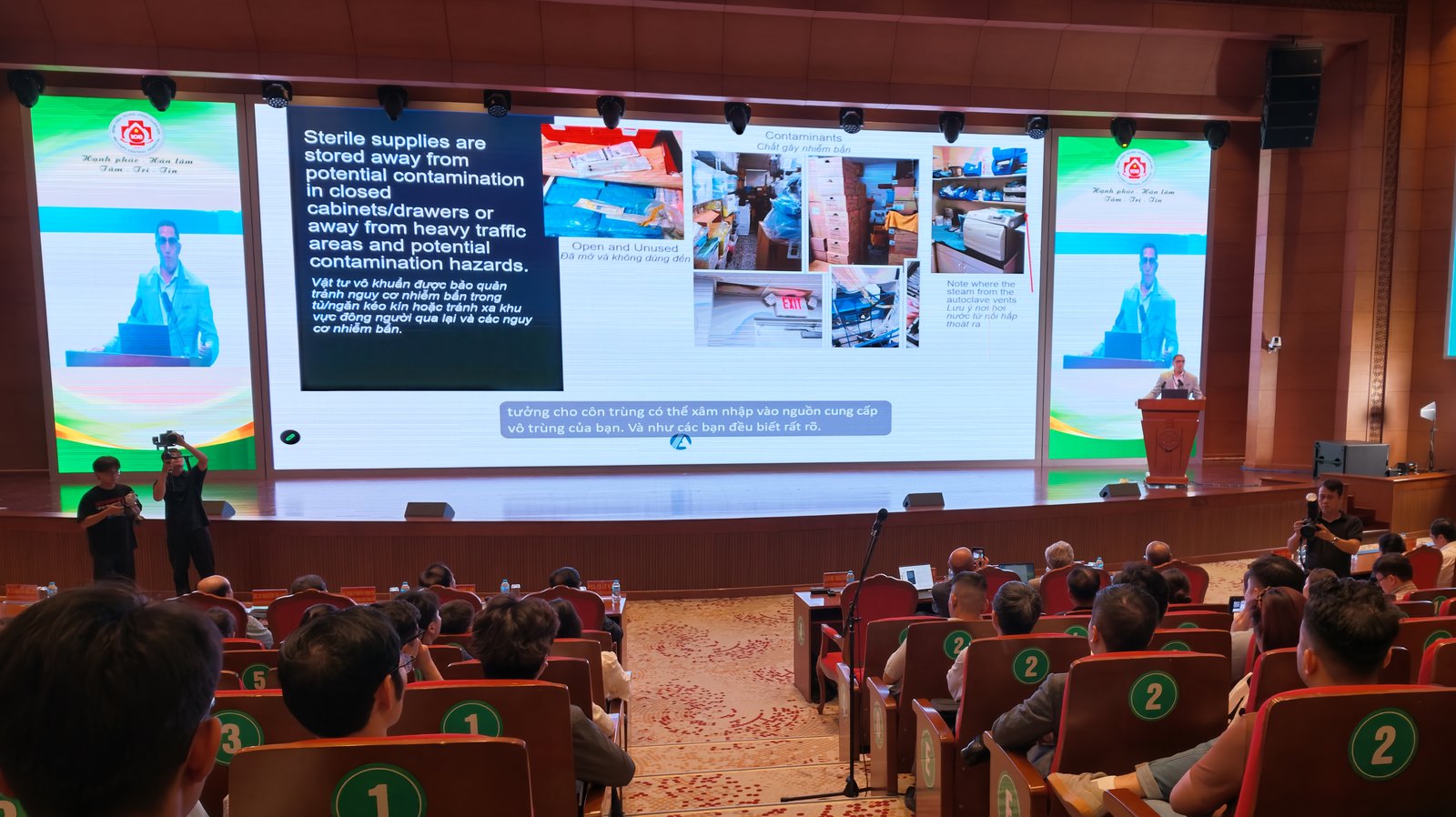 Hội trường chương trình tập huấn, trên màn hình là slide song ngữ Anh–Việt về bảo quản vật tư vô khuẩn tránh nguy cơ nhiễm bẩn